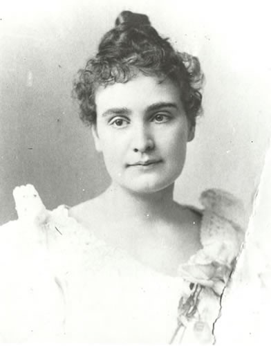 Head and shoulders portrait of Anne Sullivan, circa 1894. In this image, Anne faces the camera with a slight smile. Her head is tilted a little to her right. A long thick braid of hair appears to be curled at the top of her head and curly wisps of hair frame her face. Her light-colored dress has a wide neck with lace edging. A rose is pinned to her gown.