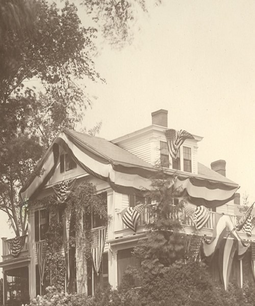 Photograph of the exterior of their three-story home in Wrentham, Massachusetts, circa 1910. The balconies on either side of the house, as well as the facade and gable, are draped with decorative material and American flags.