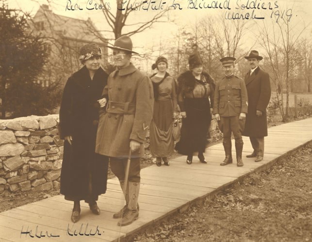 Anne, Helen, and Polly visit Evergreen, the Red Cross Institute for Blinded Soldiers in Baltimore, Maryland, March 1919. This picture shows Helen walking with a blinded soldier, her arm linked with his. Polly and Anne follow behind with a Red Cross official in uniform and another man in civilian clothes.