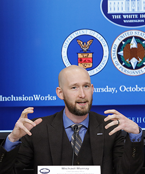 Michael Murray, wearing a suit and gesturing with both hands. Behind him the White House, Department of Labor, and US Office of Personnel Management logos are partially visible.