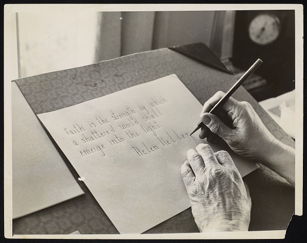 Photograph of Helen Keller's hands writing in pencil on a piece of paper that is tilted on an angled blotter. The text reads "Faith is the strength by which a shattered world shall emerge into the light. Helen Keller" Light from a window is visible in the background.
