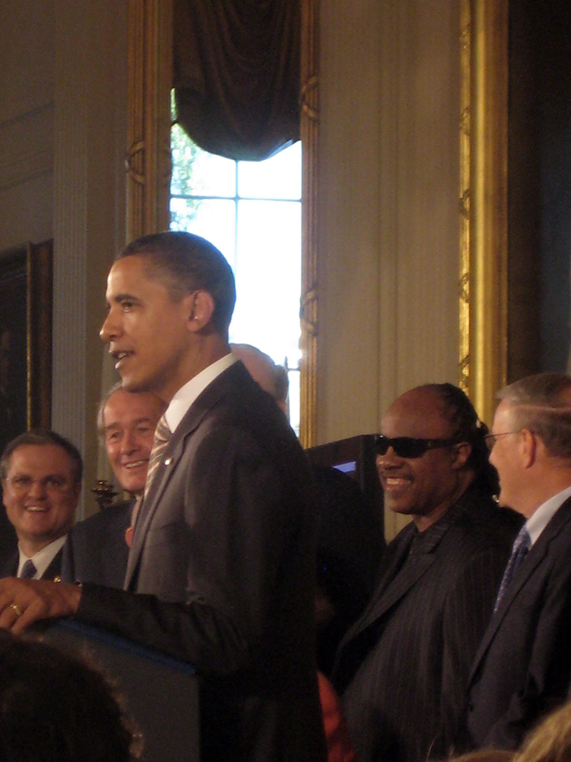 President Obama speaks at the White House during the signing of the CVAA in 2010. Stevie Wonder, Ed Markey, and others gather behind him at the podium.
