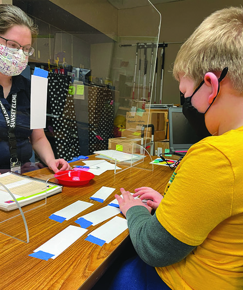 Separated by plexiglass, a White male student reads braille flashcards as his TVI looks on. She has APH's Building on Patterns on her desk. Both wear a mask. 