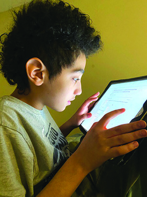 A Hispanic high school student sits at his desk using his iPad to take part in online education. 