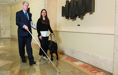 A professionally dressed man with silver hair walks with a long white cane alongside a professionally dressed female walking with a guide dog