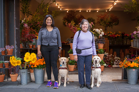 Two women stand in front of a colorful flower shop with their yellow Lab guide dogs seated at their sides.