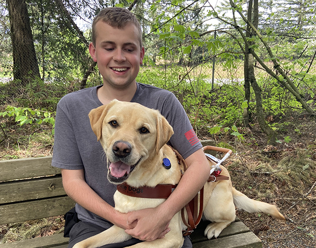 A smiling young man sits on a park bench with a smiling yellow Lab guide dog in his lap.