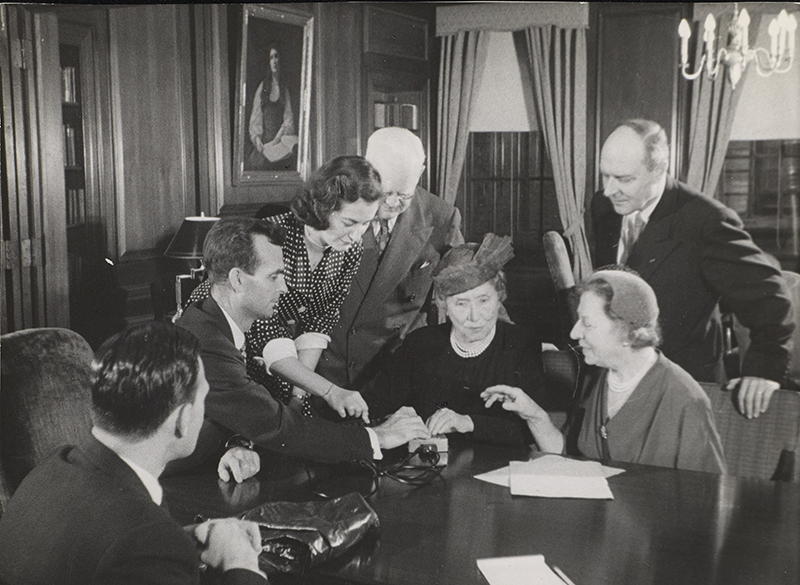 Left to right are: unidentified man, M. Robert Barnett (AFB Executive Director), Marta Sobieski, Peter J. Salmon (Executive Director, Industrial Home for the Blind, Brooklyn), Helen Keller, Polly Thomson, and Gregor Ziemer. The room has wood panelling and a painting of Helen Keller by Albert H. Munsell is visible in the background. Image from documentary Helen Keller in Her Story.
