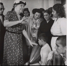 A crowd surrounds Helen Keller as she gestures. She stands in front of two boys who are blind, one of whom is holding a musical instrument. Egypt, 1952.