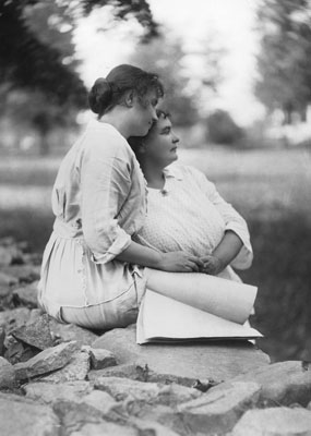 Helen Keller sitting on a stone wall with a braille book on her lap while Anne Sullivan Macy leans against the wall, 1904.