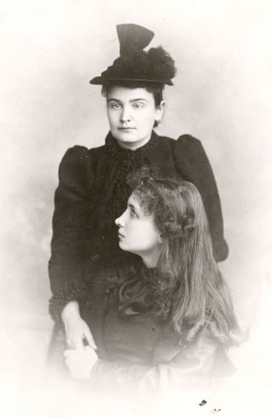 Helen Keller is holding Anne Sullivan's hand. Helen is seated in profile in front of Anne, who stands facing the camera at a slight angle. Helen's curly bangs frame her face and her long hair flows over her shoulders. Anne is dressed in dark clothing and hat. Circa 1893.