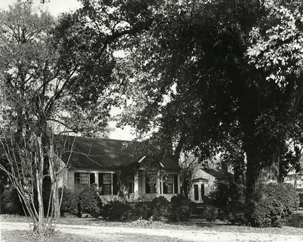This is the main house on the Keller family's Ivy Green estate. This picture shows a one-story house surrounded by big, round shrubs and tall, leafy trees. It is a sunny day, but the house is in the shade of all the plants. The house has white wood shingles and lots of windows with dark shutters.