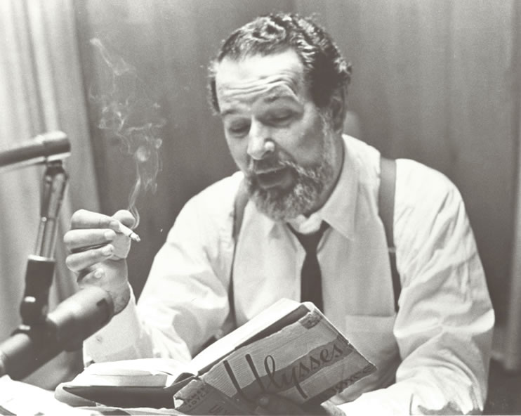 Talking Book narrator Alexander Scourby is seen from the waist up, sitting in a recording studio narrating Ulysses by James Joyce. He holds a lit cigarette in his right hand and the open book in his left. Photograph circa 1970s. Talking Book Archives, American Foundation for the Blind.