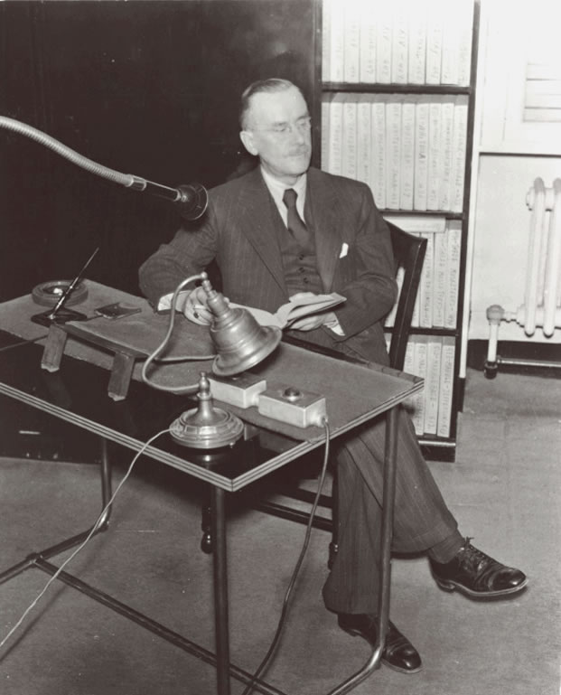 Author Thomas Mann recording his book Buddenbrooks in 1941. Mann is seated sideways to a table. He holds a cigarette and his book. He wears a pin-striped three-piece suit with a tie. An angled lamp sits on the table and a microphone with a long-coiled metal neck faces him. A shelf of containers of Talking Book records are behind him. Talking Book Archives, American Foundation for the Blind.