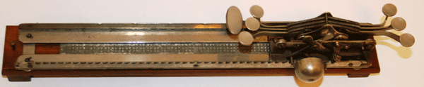 This wood and metal portable braillewriter is in the shape of a ruler. It is 13 inches wide by 3 inches high and 3 inches deep. The long side of the braillewriter faces the user, who depresses keys that are on the right-hand side. These keys punch holes in a metal grid of braille dot cells.