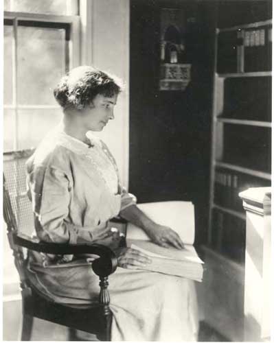 Helen Keller seated in a library in front of a window, reading a braille book with her left hand