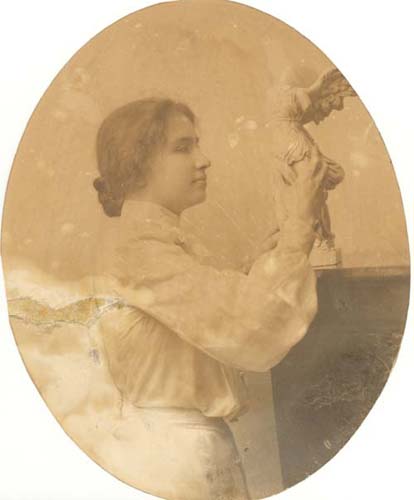 Helen Keller standing at a mantle, her face in profile, exploring a white statue of a winged figure.