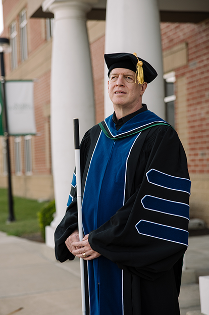 Kirk Adams, standing with his cane, wearing academic cap and gown.