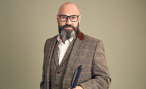 Dave Steele, wearing a gray suit with a white shirt and no tie. His head is shaved and he has a salt-and-pepper beard. He is wearing dark-framed glasses and holding a white cane.