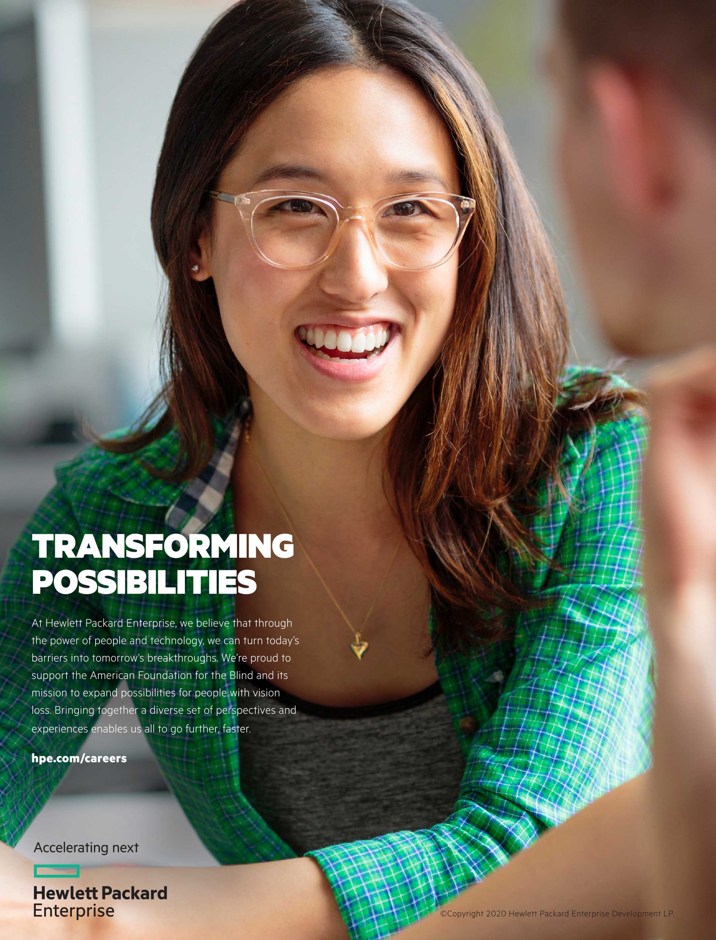 Image: Full-page close up of a young, smiling Asian woman with long brown hair wearing glasses and a green and blue button-down shirt, seated at a table and talking to someone outside of the image.