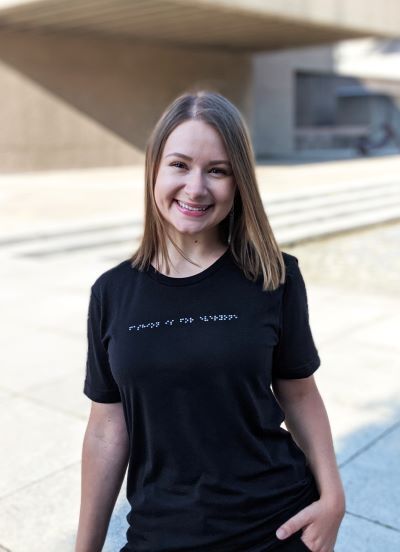 Alexa Jovanovic, founder of Aille Design, is smiling and wearing a black t-shirt with Swarovski Crystal beading that reads “Fashion is for everyone.” The t-shirt with tactile Braille is available for purchase at www.ailledesign.com.