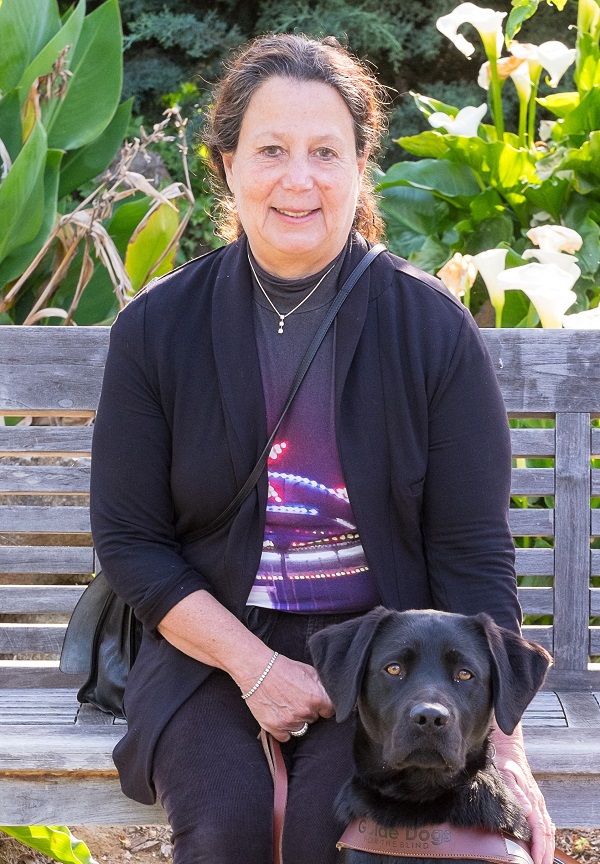 Janni Lehrer-Stein sitting on a park bench. Next to her is her dog guide Shilo, a black labrador retriever. 