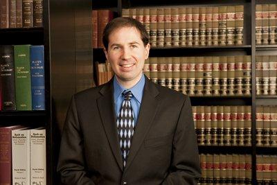 Tim Elder, attorney, wearing a suit and tie, smiling. He stands in front of a shelf full of legal books.