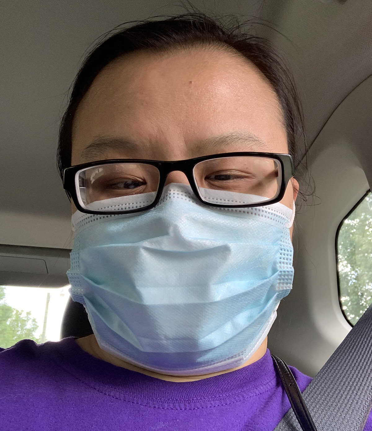 An Asian woman wears glasses and a mask as a passenger in a car.