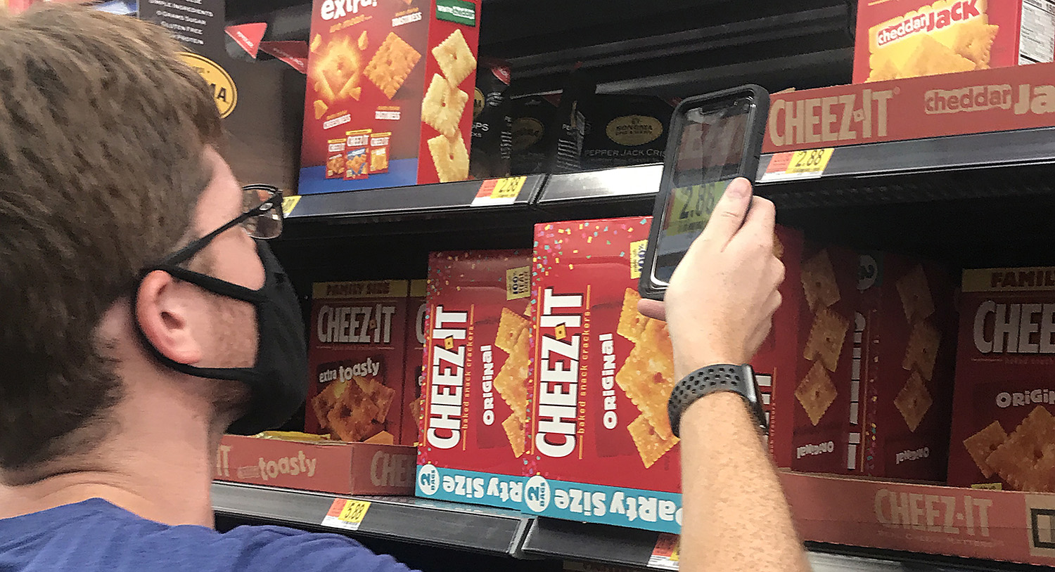 A White man wearing a mask uses an app on his smartphone to read the price of an item on a grocery store shelf. 