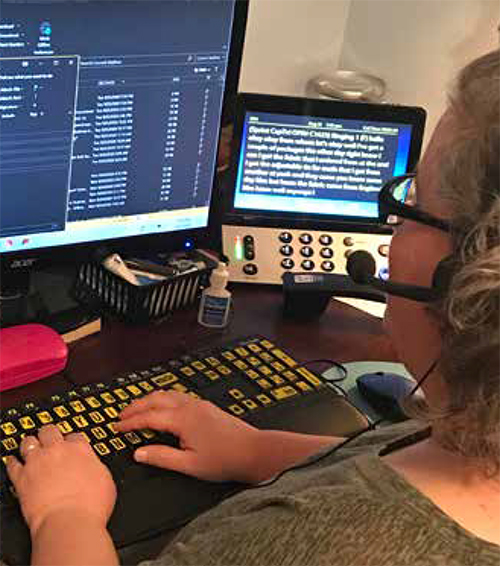 An older White woman wearing a headset types on a high-contrast keyboard that has yellow keys with black text. She is using reverse contrast on the computer screen with white text on a dark-blue background. Her video phone is to the right and also displays white text on a dark blue background.