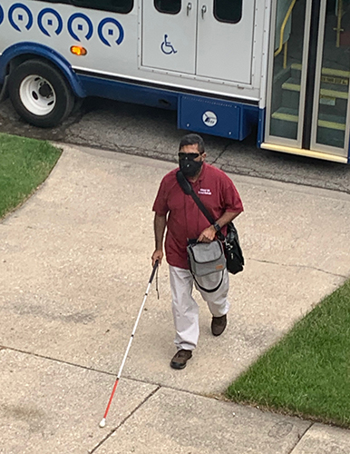 An Indian man wearing a mask uses a white cane after leaving a paratransit van. 