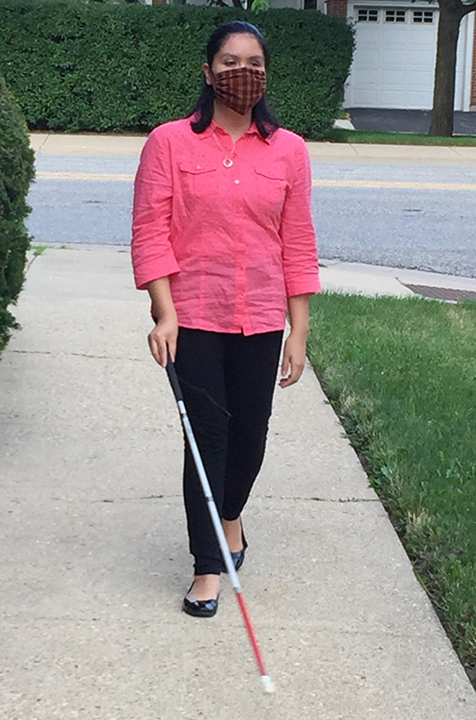 A woman walks with her cane on a residential sidewalk. She is wearing a mask, a pink shirt, and black pants. 