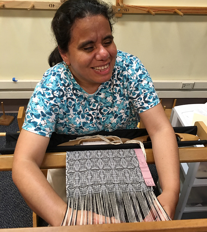 A smiling Latina woman is weaving on a loom. The material she is weaving has an intricate black and white design.