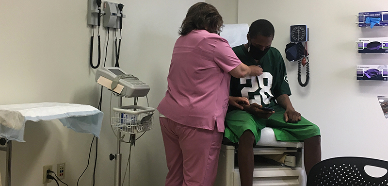 A young Black man wearing a mask is examined by a nurse in a doctor’s office.