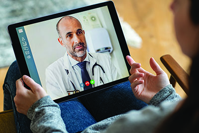 A telemedicine session takes place between a doctor, visible on a tablet screen, and a patient who is at home holding the device.