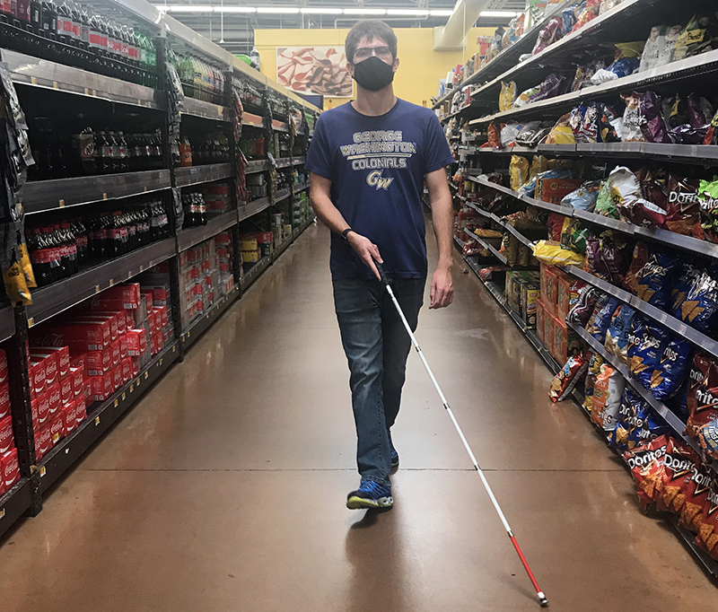 A young White man wearing a mask walks down a supermarket aisle with his white cane.