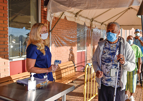 A White woman wearing a mask holds a thermometer and addresses a Black man wearing a mask and holding a white cane. He stands at the front of a line of people in a covered outdoor area. 