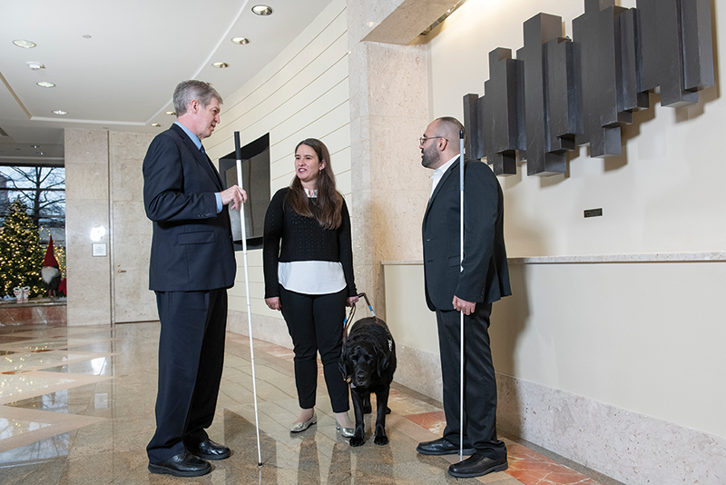 Kirk Adams holds his white cane and stands in a large lobby with a young White woman with her guide dog and a young Middle Eastern man with his white cane.