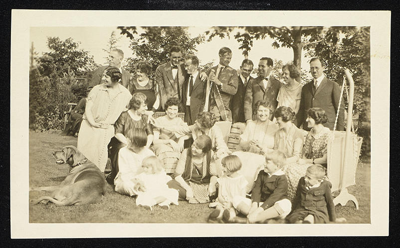 Helen Keller, Polly Thomson, and Anne Sullivan Macy? are sitting in the garden with a group of deaf friends at their home in Forest Hills. Men, women and children are smiling and many appear to be looking to their left. The children sit infront of the adults on the grass, the girls have bobbed haircuts. Thomson sits in the middle row with Macy next to her. Keller is the fifth person from the left. Men and women are standing behind them. Keller shares a garden swing chair with three other women.