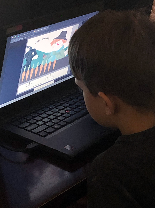  A white elementary boy is at his laptop using the program Origio Math. He is leaning into the screen to select the number of carrots to complete the math problem 9 - ___ = 6. Not visible on the screen are his classroom teacher and paraprofessional who are working with him.