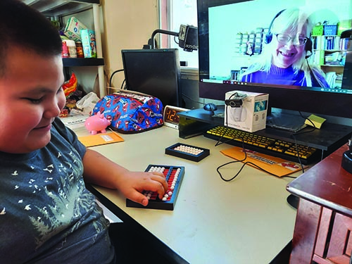 An elementary Native American boy smiles as he uses a Cranmer abacus at home as his smiling TVI watches via Zoom. 