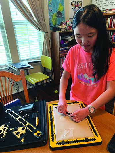 In her homeschool classroom a Chinese-American girl uses the TactiPad Drawing Board and compass to draw a circle. To her right, the case that holds the TactiPad Drawing Board and tools is visible. 