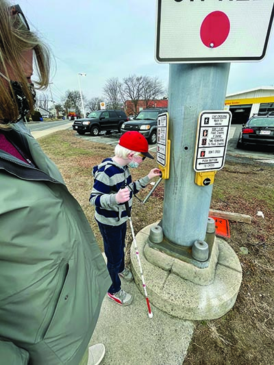At an intersection a white elementary boy with albinism wearing a mask presses the signal button with his left hand. His cane is in his right hand. His white female O&M specialist who is also wearing a mask stands nearby. 