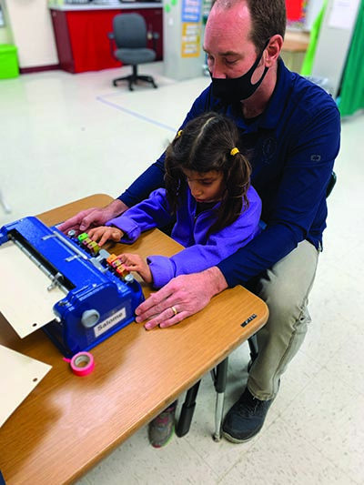 A White elementary girl writes on a Perkins braillewriter as her White male TVI wearing a mask sits behind her with his arms around her ready to provide support. 