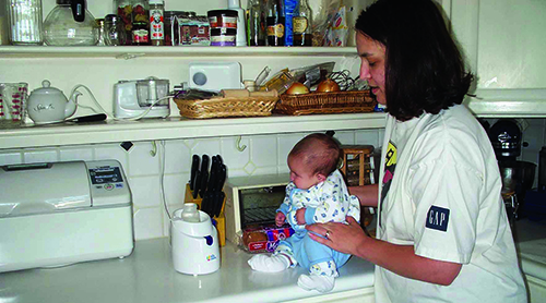 A White infant waits for his bottle to warm as he sits on the counter. His mother holds him in a sitting position as they both listen for the bell to signal the bottle is warm. 