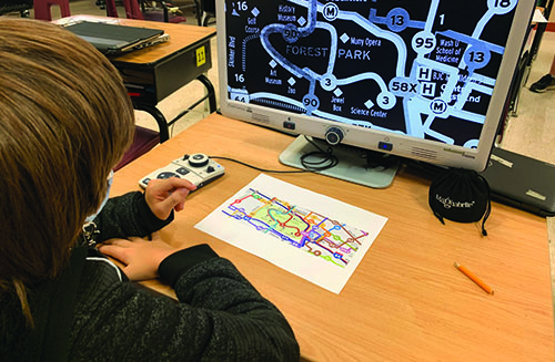 A White elementary boy uses a video magnifier to enlarge a transit map. He has chosen to use white on black for higher contrast. 