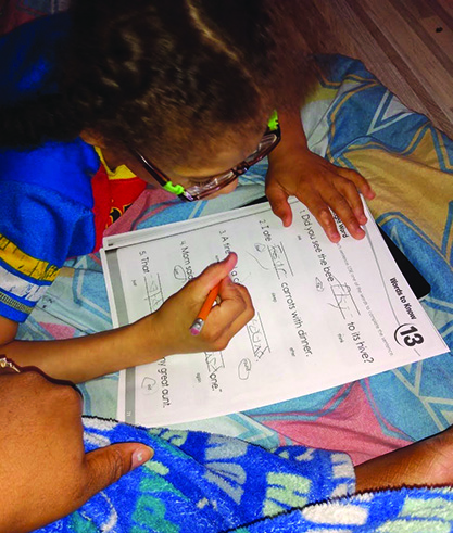 A Black elementary school-aged boy sits on his bed completing a language arts worksheet. 