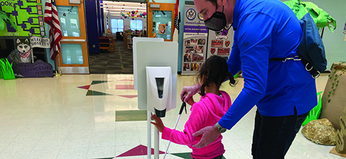 In a school hallway, a White elementary school-aged girl and her White O&M specialist use her cane to find a hand sanitizing station. His hand is on top of hers and he wears a mask. 