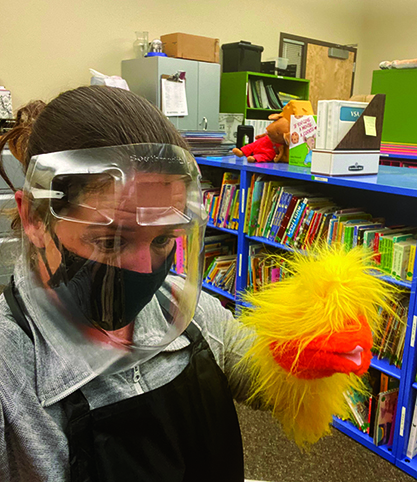 A White TVI wearing a black mask and face shield stands in a classroom. She has a fuzzy yellow duck puppet on her arm. 
