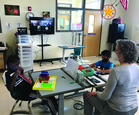 In a classroom, a White teacher wearing a mask sits with two elementary students who are separated by plexiglass and each wearing a mask. Both boys, one African American and one Hispanic, use math manipulatives on APH work trays. Other classmates are on a computer screen mounted to the wall. 